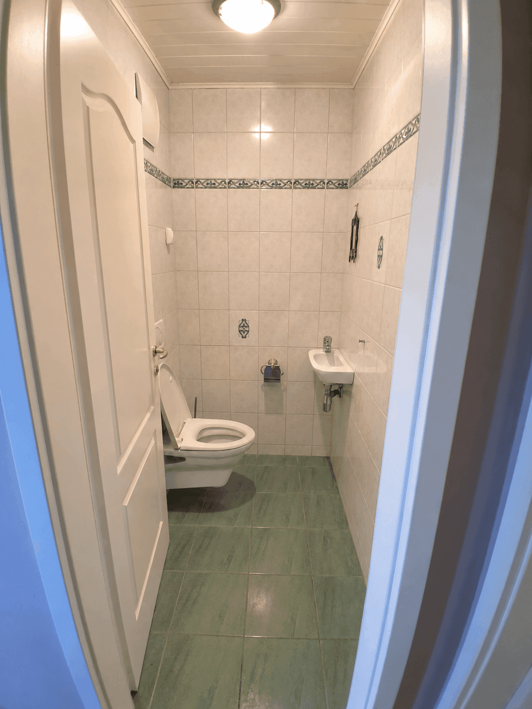 Enges Gäste-WC mit Wand-WC und kleinem Waschbecken, gefliest in Grün und Weiß.