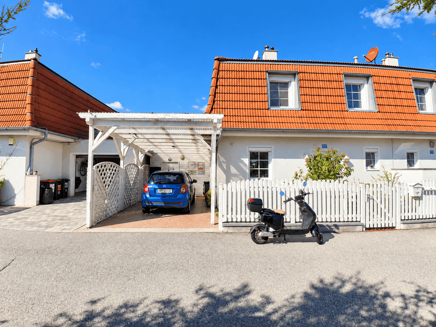 Zwei Wohnhäuser mit roten Ziegeldächern, weißer Fassade, Carport und einer weißen Zaunanlage an der Straße.