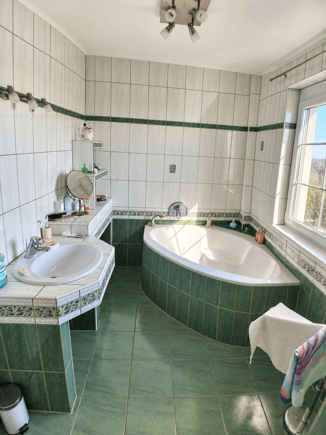 Helles Badezimmer mit Eckbadewanne, Waschbecken und Fenster, gefliest in Grün und Weiß.