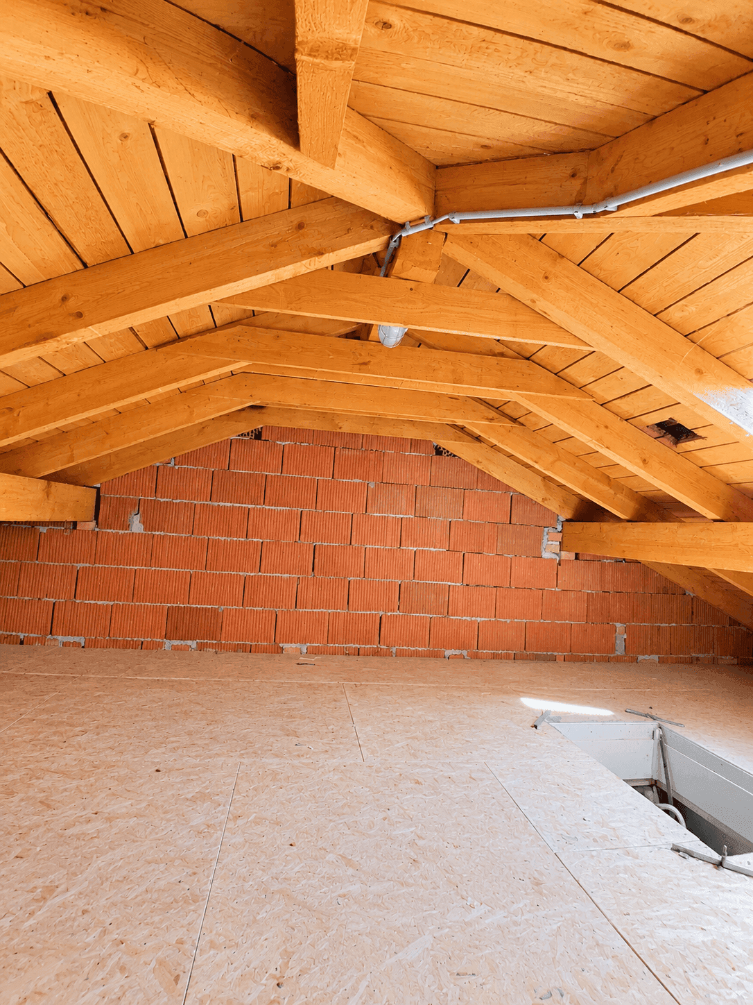 Dachboden mit Holzbalken, unverputzter Ziegelwand und OSB-Platten als Bodenbelag.