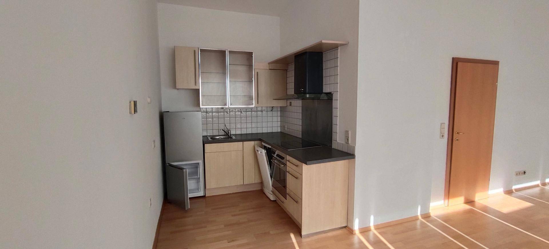 Kleine Küchenzeile mit hellen Schränken, Kühlschrank und Kochfeld in einem offenen Raum.
