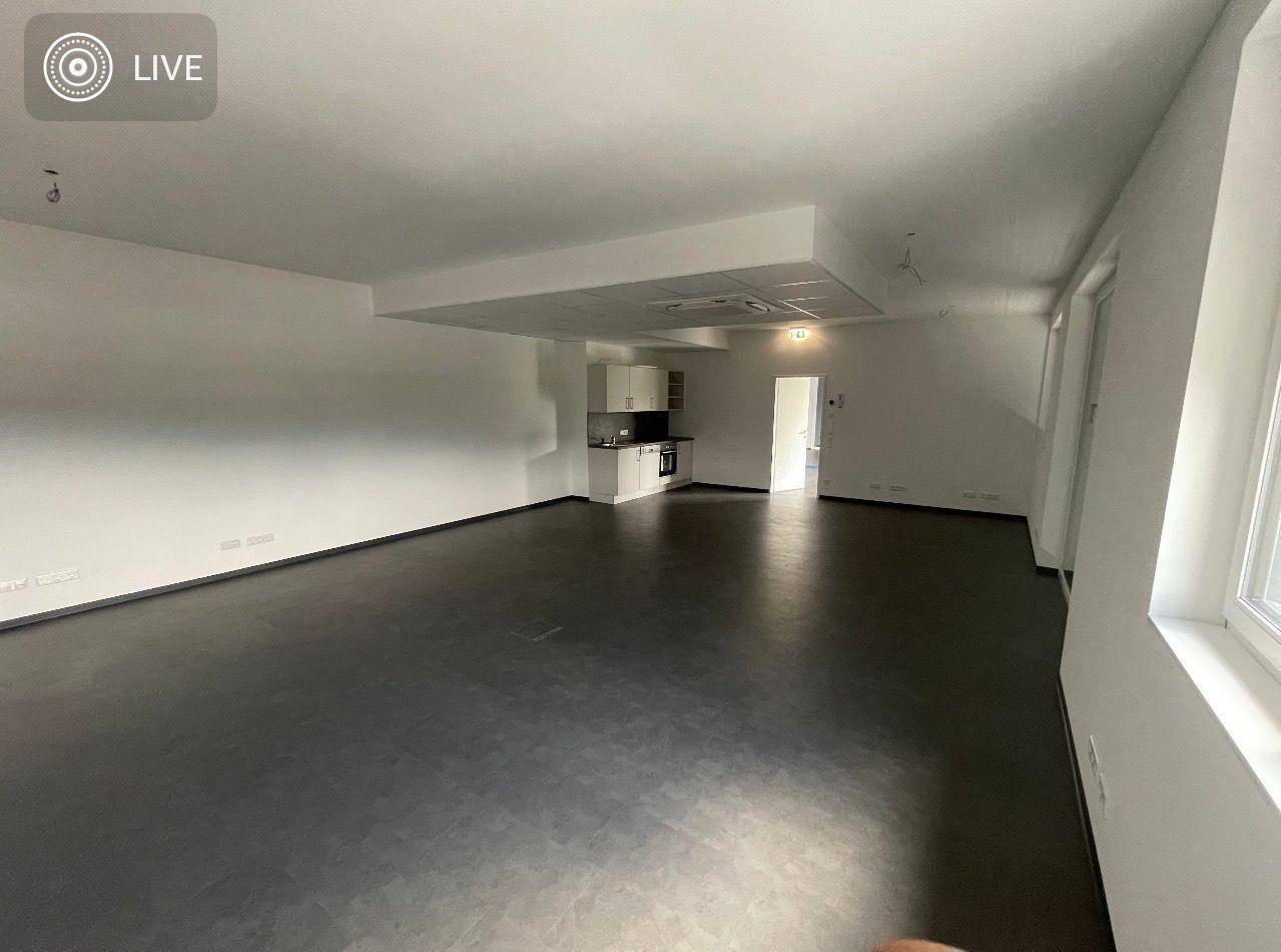 Heller, offener Büroraum mit dunklem Boden und Blick auf eine kleine Teeküche im Hintergrund.