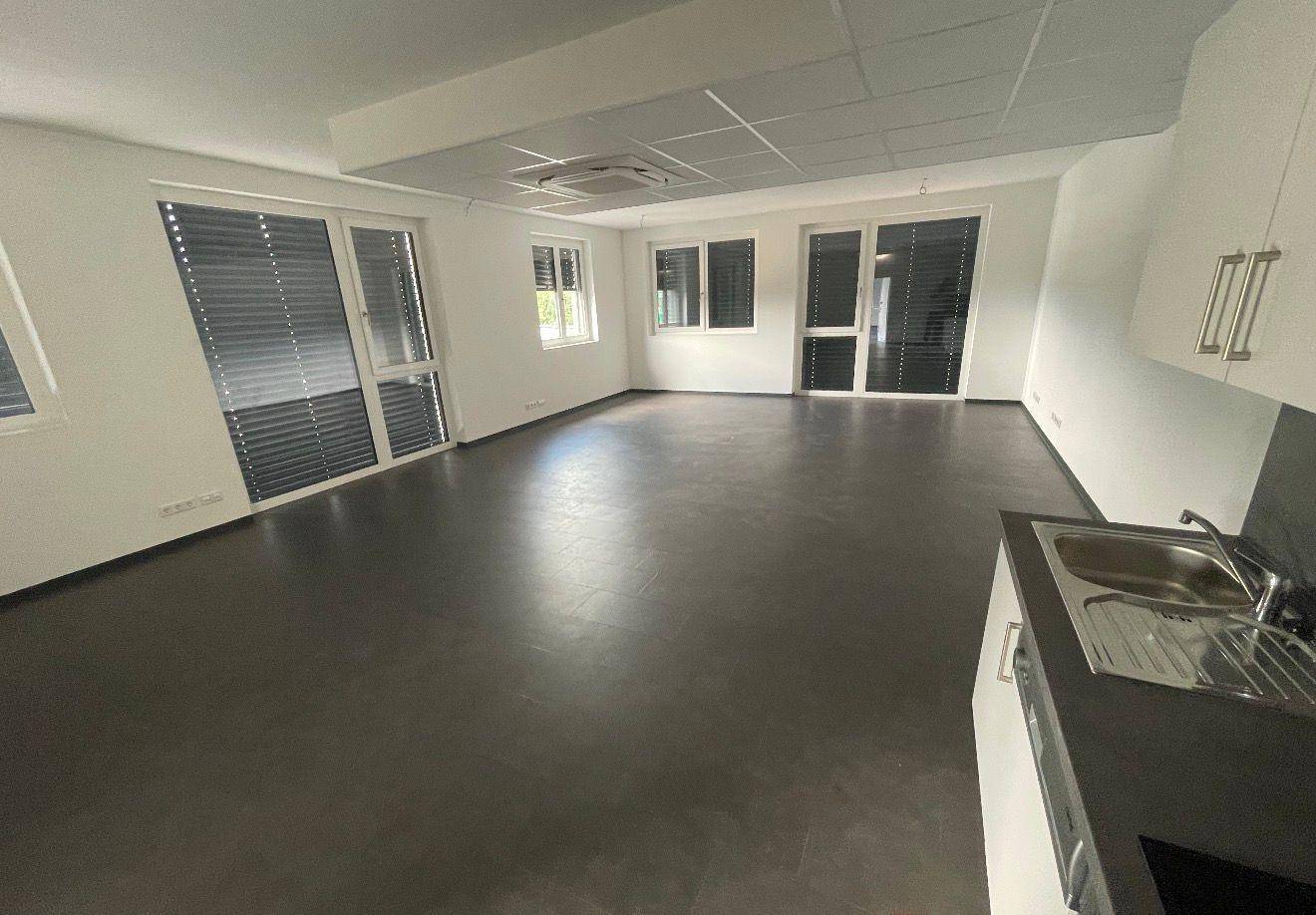 Geräumiger Büroraum mit dunklem Vinylboden, großen Fenstern und integrierter Teeküche.