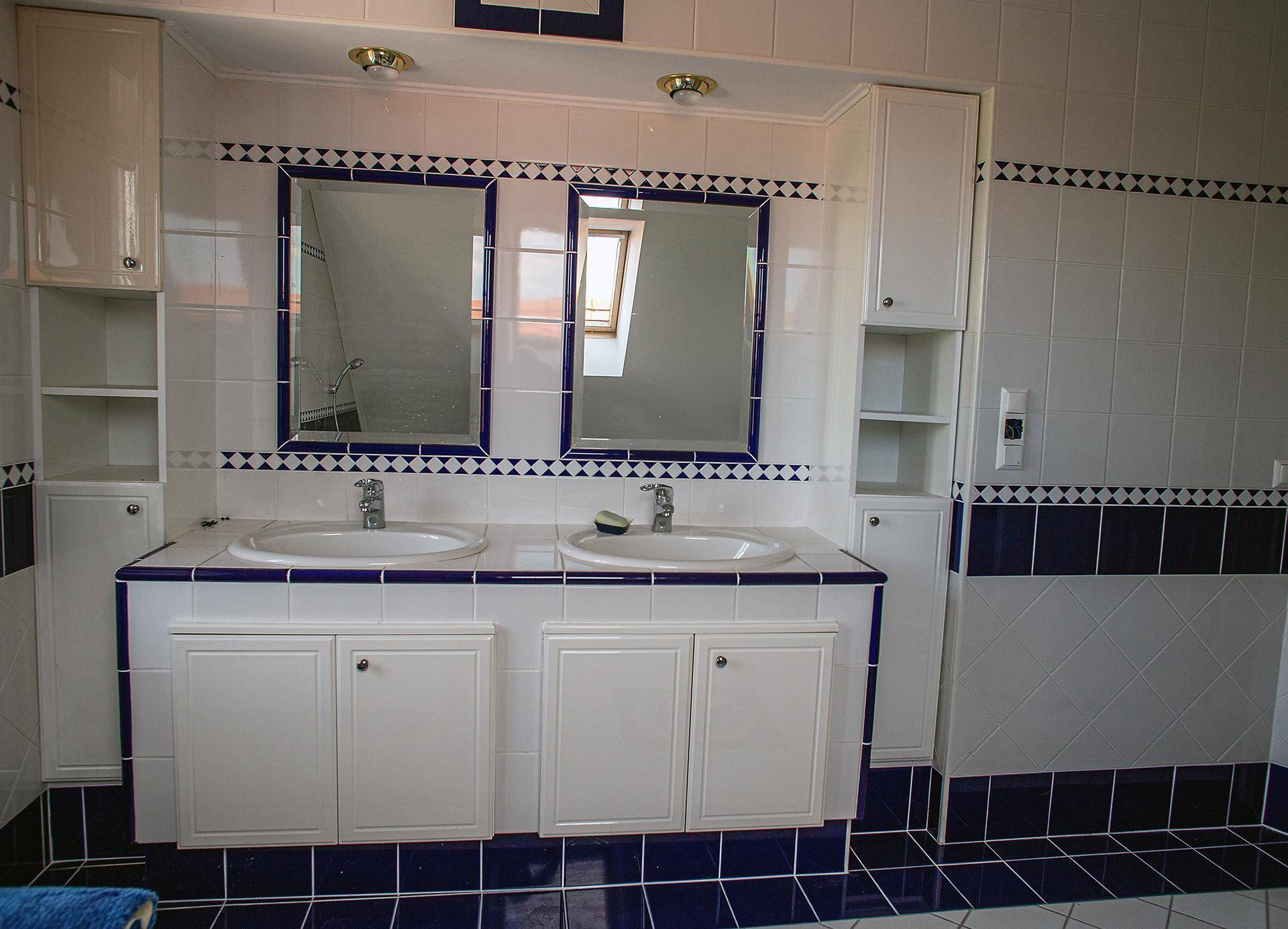 Badezimmer mit Doppelwaschbecken, weiß-blauen Fliesen und Spiegeln, die ein Dachfenster reflektieren.