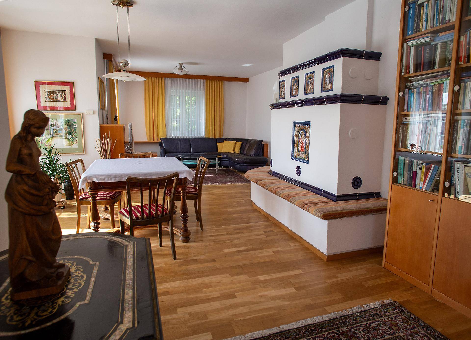 Geräumiger Wohn-Essbereich mit Holzboden, Kachelofen und einem Bücherregal, ideal zum Entspannen und Lesen.