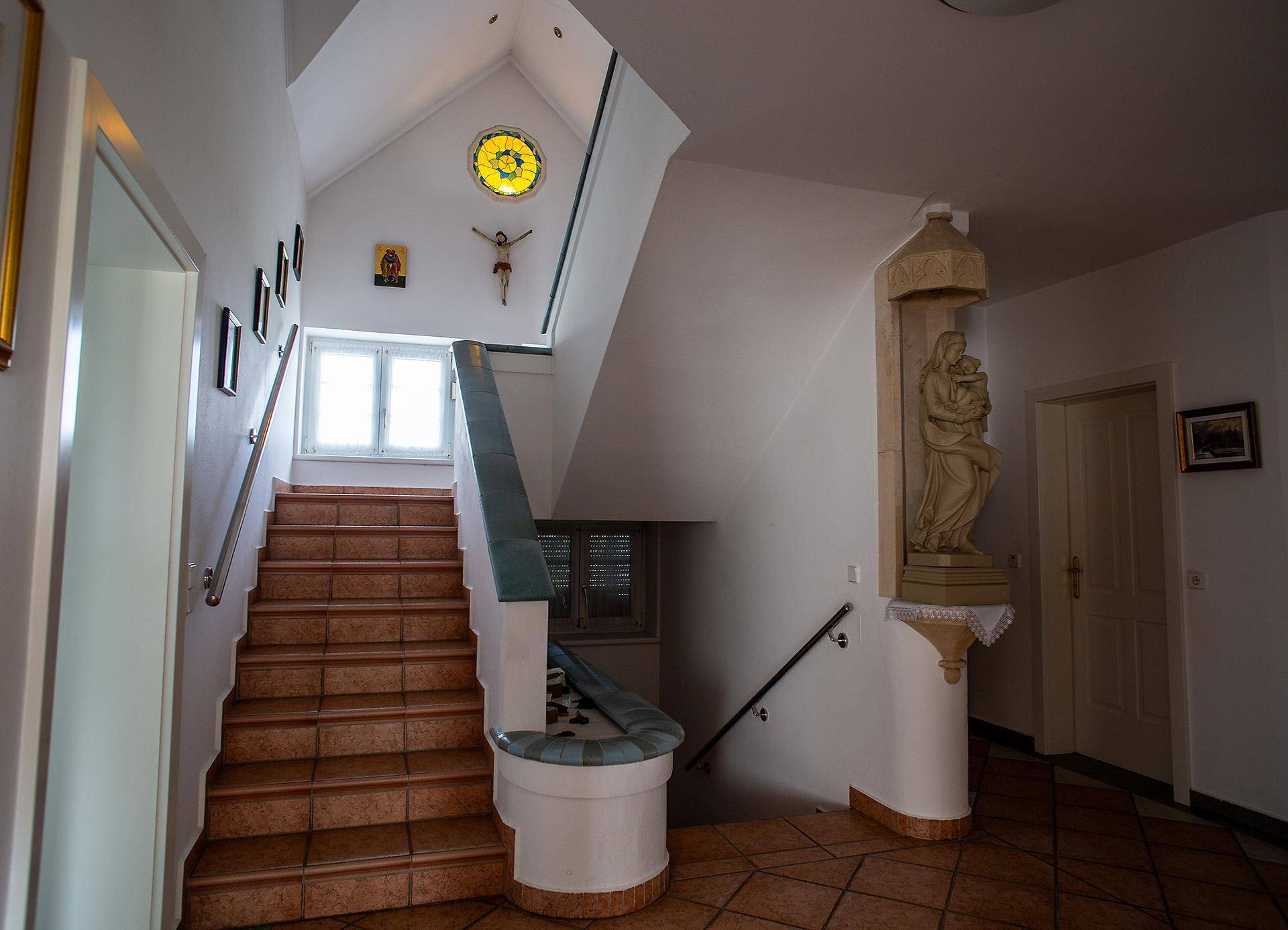 Heller Flur mit gefliester Treppe, religiösen Dekorationen und einem Buntglasfenster im oberen Bereich.