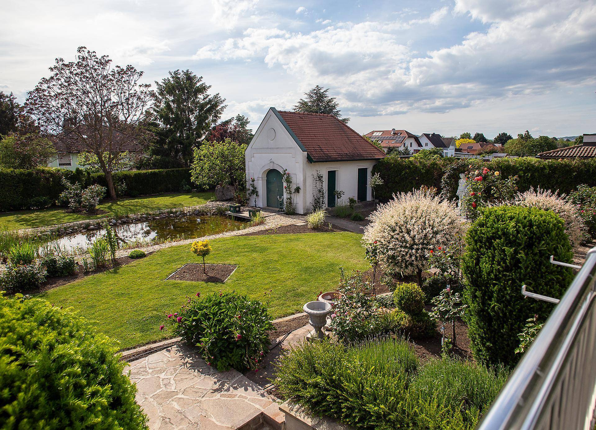 Erhöhter Blick auf den gepflegten Garten mit Teich, Nebengebäude und üppiger Vegetation von oben.