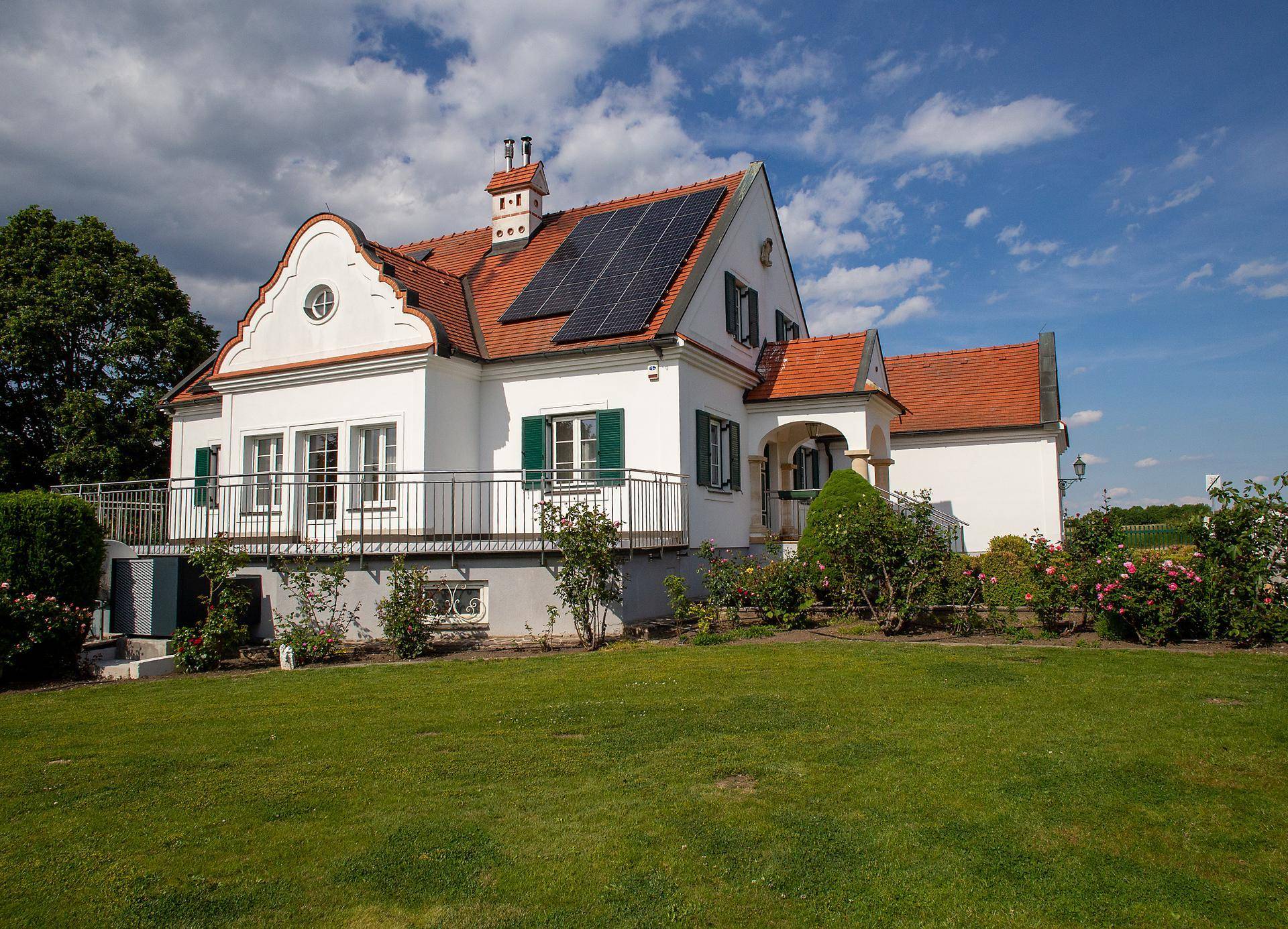 Seitenansicht des Hauses mit Solaranlage auf dem Dach und einem Balkon über dem grünen Gartenbereich.