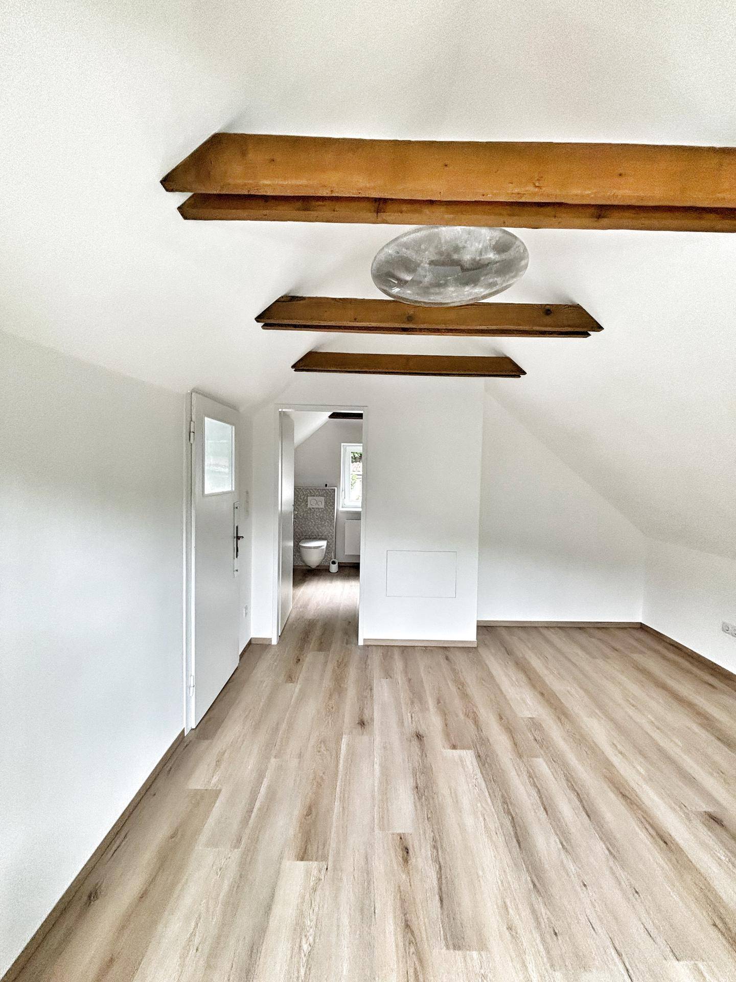 Geräumiges Dachzimmer mit sichtbaren Holzbalken und hellem Laminatboden, mit Zugang zu einem Badezimmer.