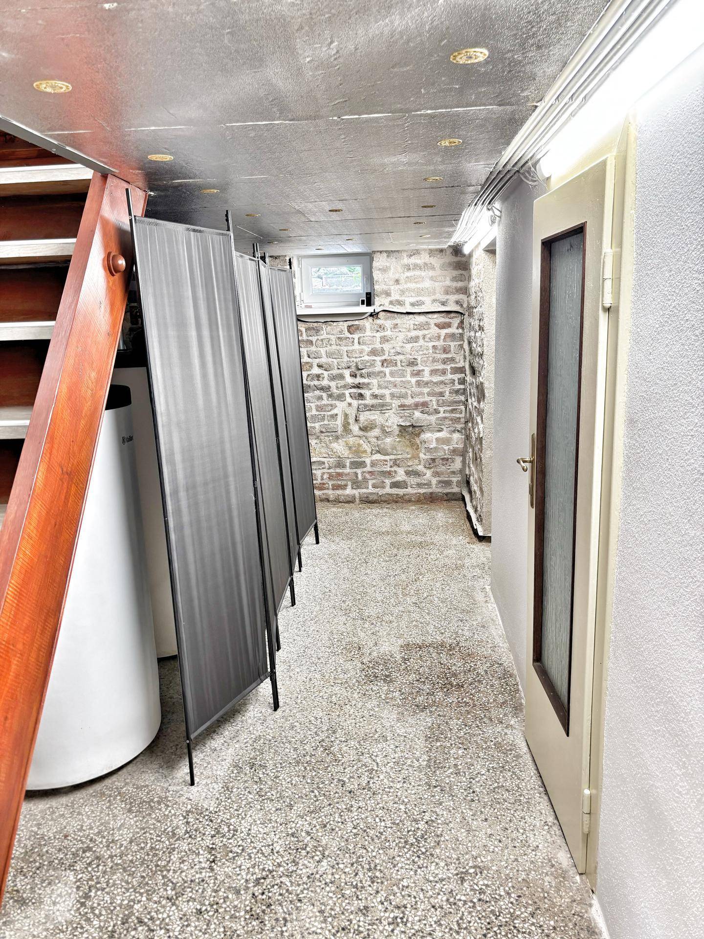 Kellerraum mit freiliegender Ziegelwand, Betonboden und einem Warmwasserspeicher, als Abstellraum nutzbar.