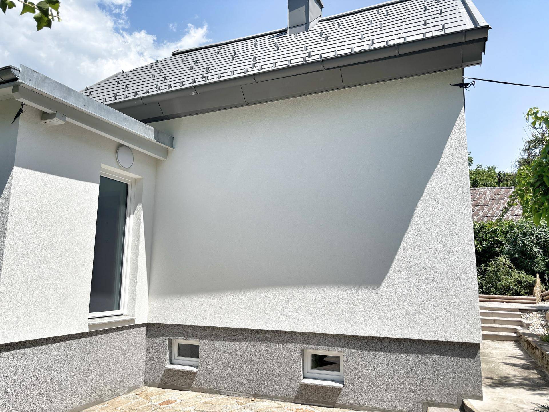 Hausfassade mit weißem Putz und grauer Sockelzone, umgeben von einem Steinfliesenweg.