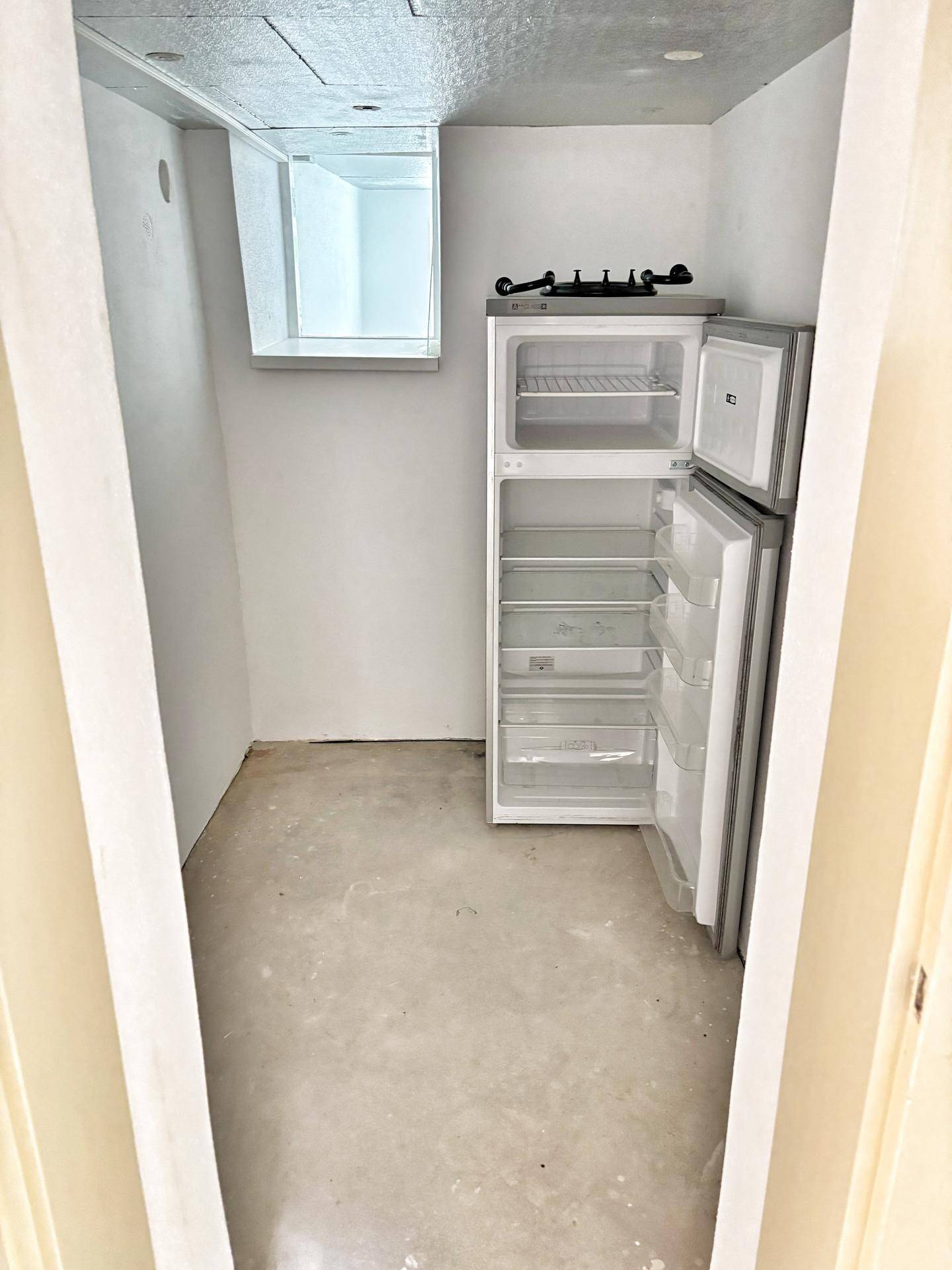 Praktischer Hauswirtschaftsraum mit Betonboden, Kühlschrank und kleinem Fenster, ideal für Vorräte.