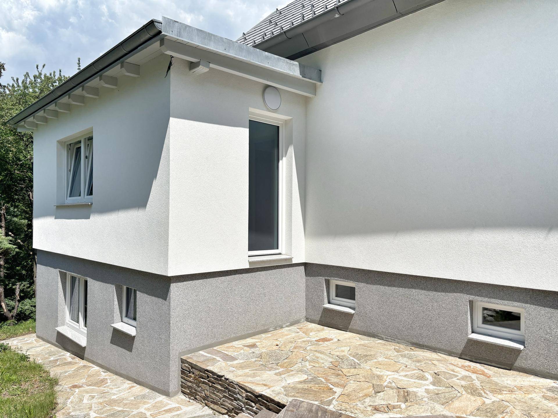 Seitenansicht des Hauses mit weißer Fassade, grauer Sockelzone und Fenstern zum Garten.