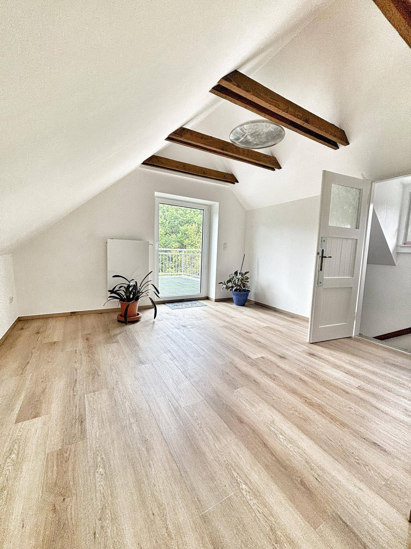 Großes Dachzimmer mit Holzbalken, hellem Boden und Zugang zu einem Balkon, ideal als Schlaf- oder Arbeitszimmer.