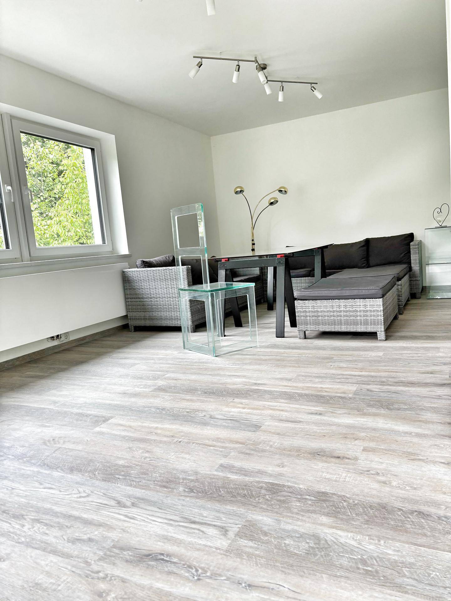 Geräumiger Wohnbereich mit hellem Laminatboden, großen Fenstern und moderner Einrichtung.