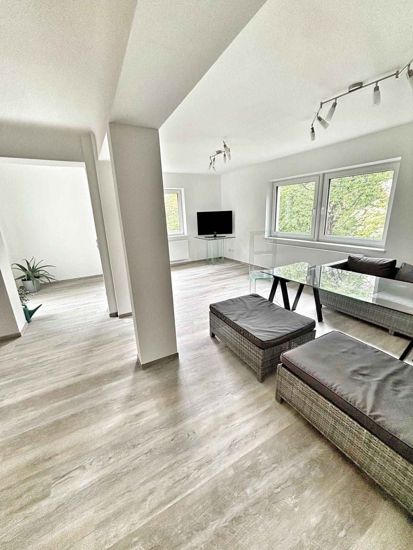Heller Wohnbereich mit großen Fenstern, modernem Laminatboden und schlichten Möbeln.
