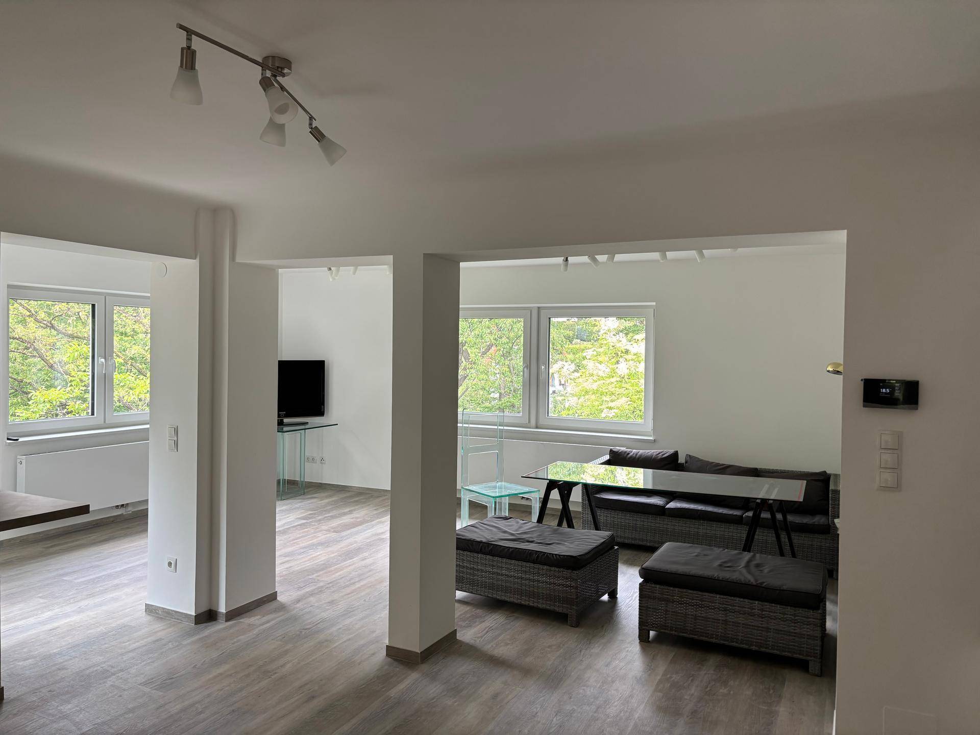 Heller Wohnbereich mit großen Fenstern, modernem Laminatboden und schlichten Möbeln.