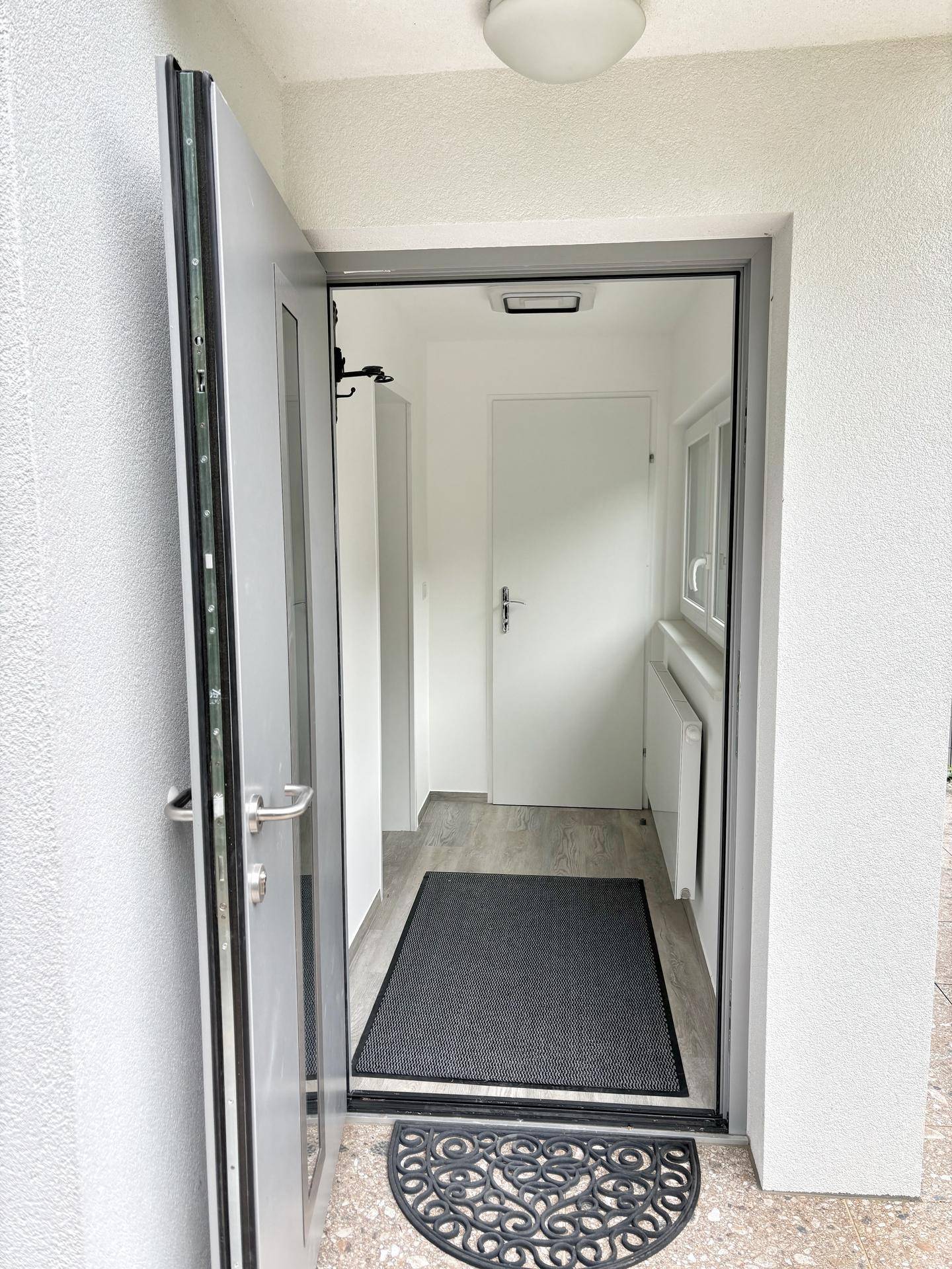 Heller Eingangsbereich mit moderner grauer Tür, hellem Boden und weiß gestrichenen Wänden.