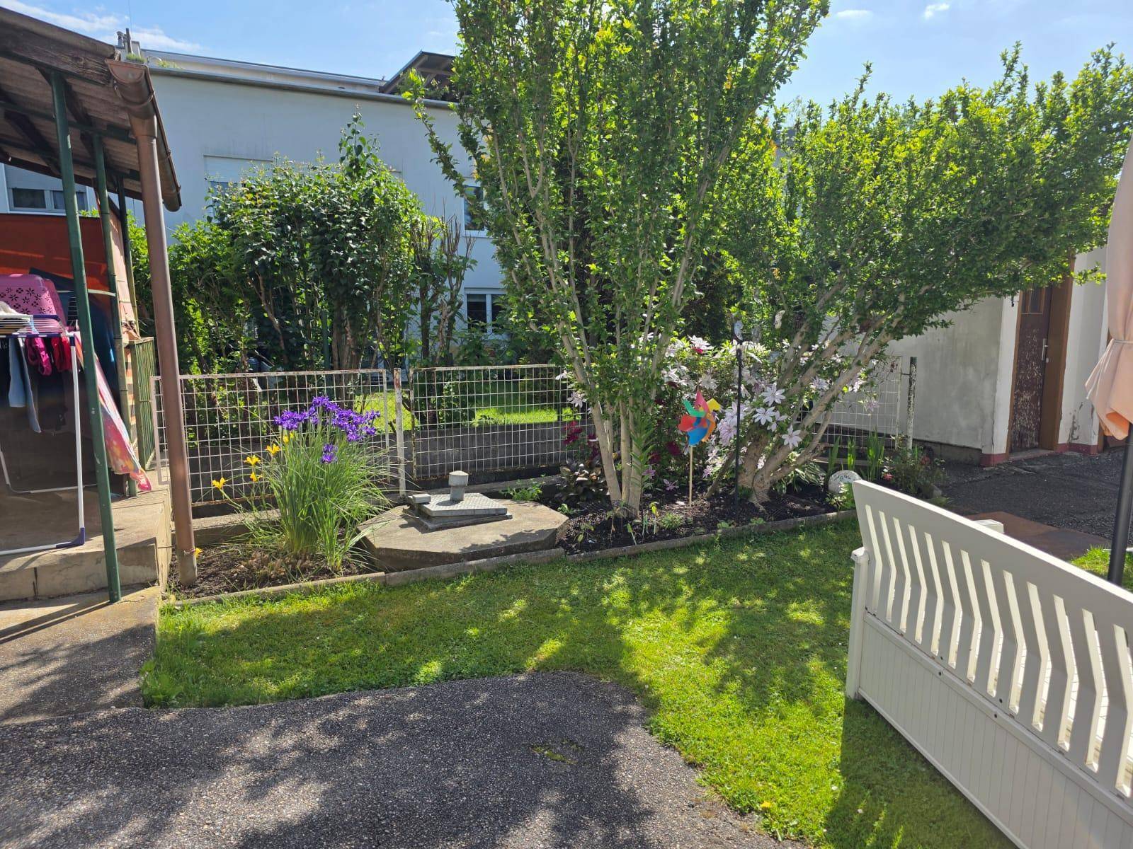 Bepflanzter Gartenbereich mit Blumen, einer Gartenbank und einem dekorativen Brunnen.