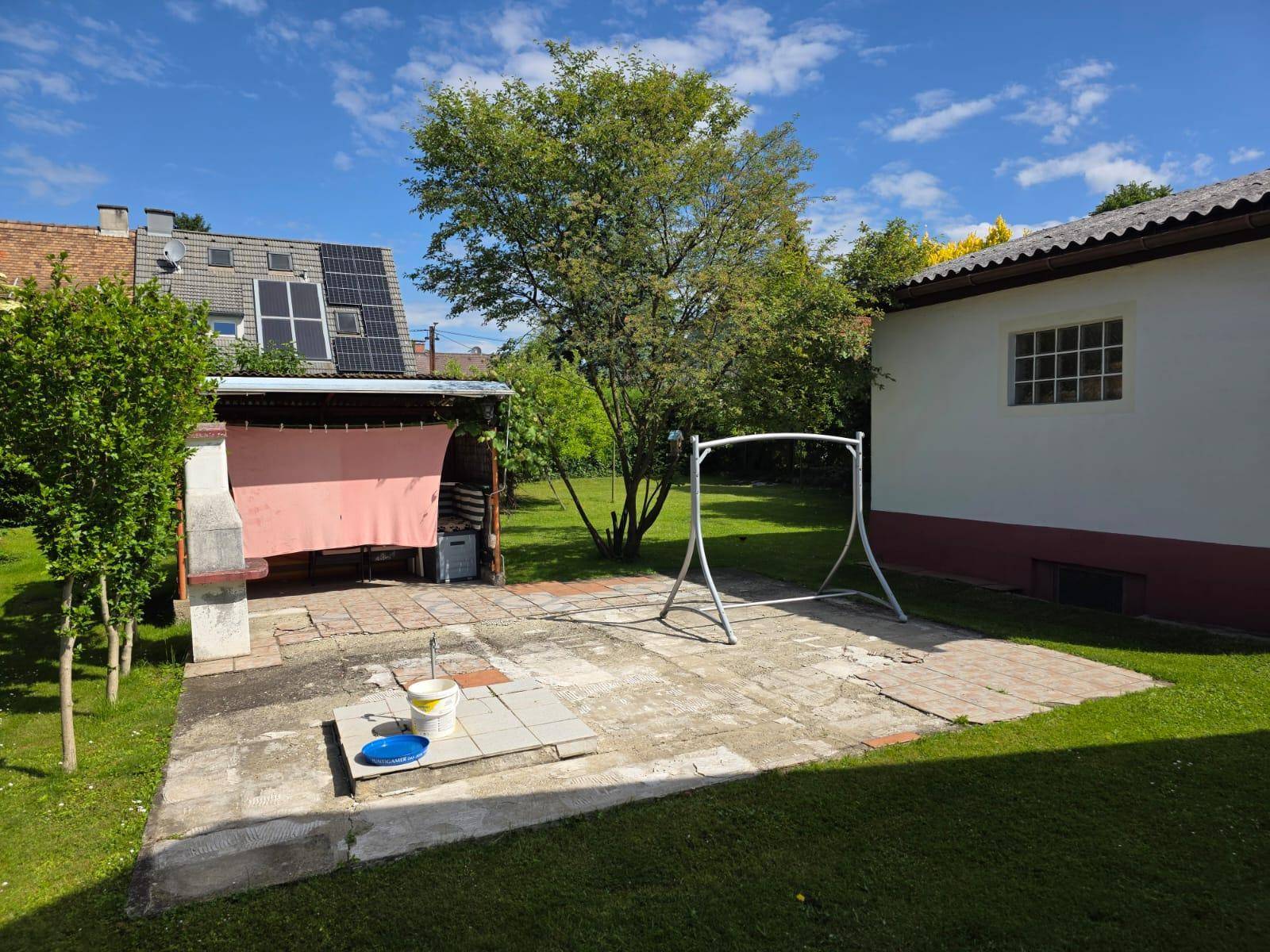 Große gepflasterte Terrasse mit Grillplatz und Schaukelgestell, umgeben von Rasenflächen.