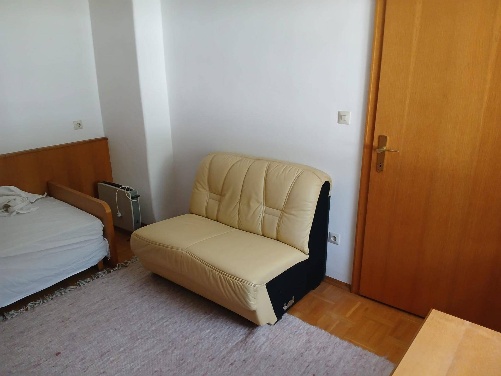 Helles Schlafzimmer mit beigem Sofa, Holztür und teilweise sichtbarem Parkettboden.