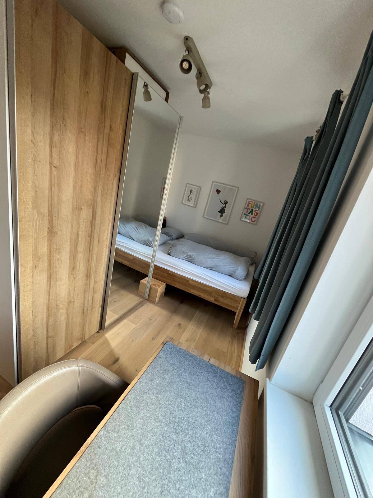 Schlafzimmer mit Holzbett, Spiegelschrank und Holzboden, gut beleuchtet durch das Fenster.