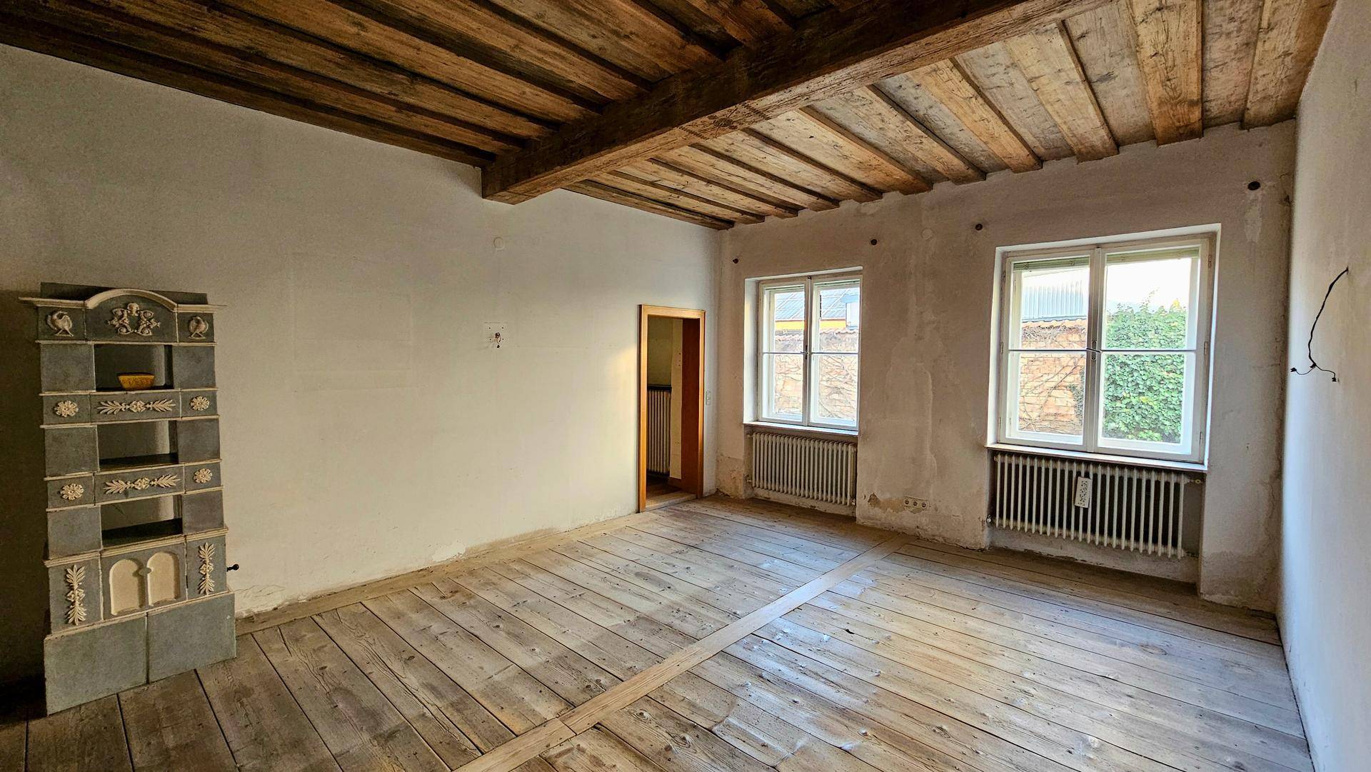 Geräumiger Raum mit Holzdielenboden, freiliegenden Deckenbalken und einem Kachelofen.
