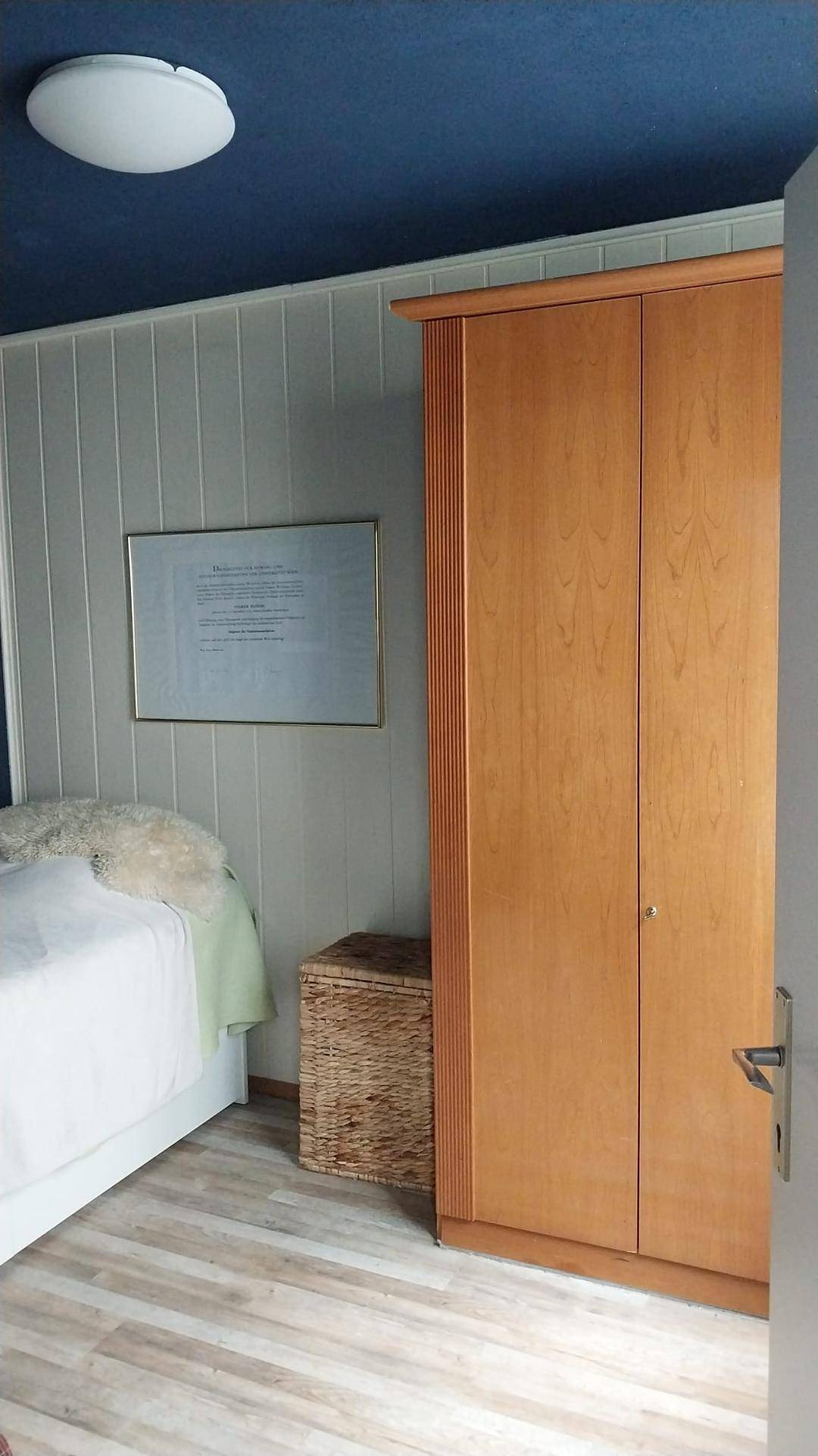 Schlafzimmer mit Holzkleiderschrank, Bett und blauer Decke sowie hellen Wänden.