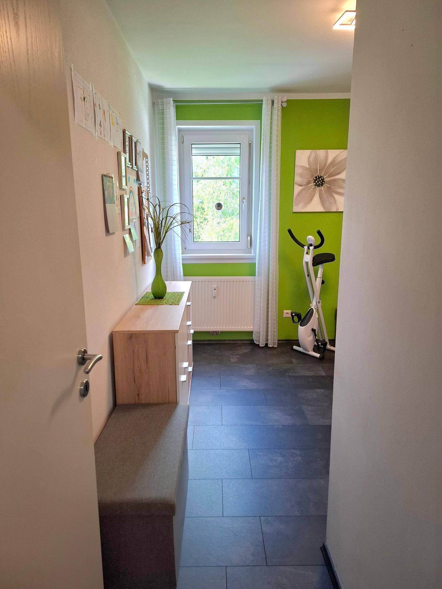 Helles Zimmer mit grünen Wänden, einem Fenster und einem Heimtrainer.