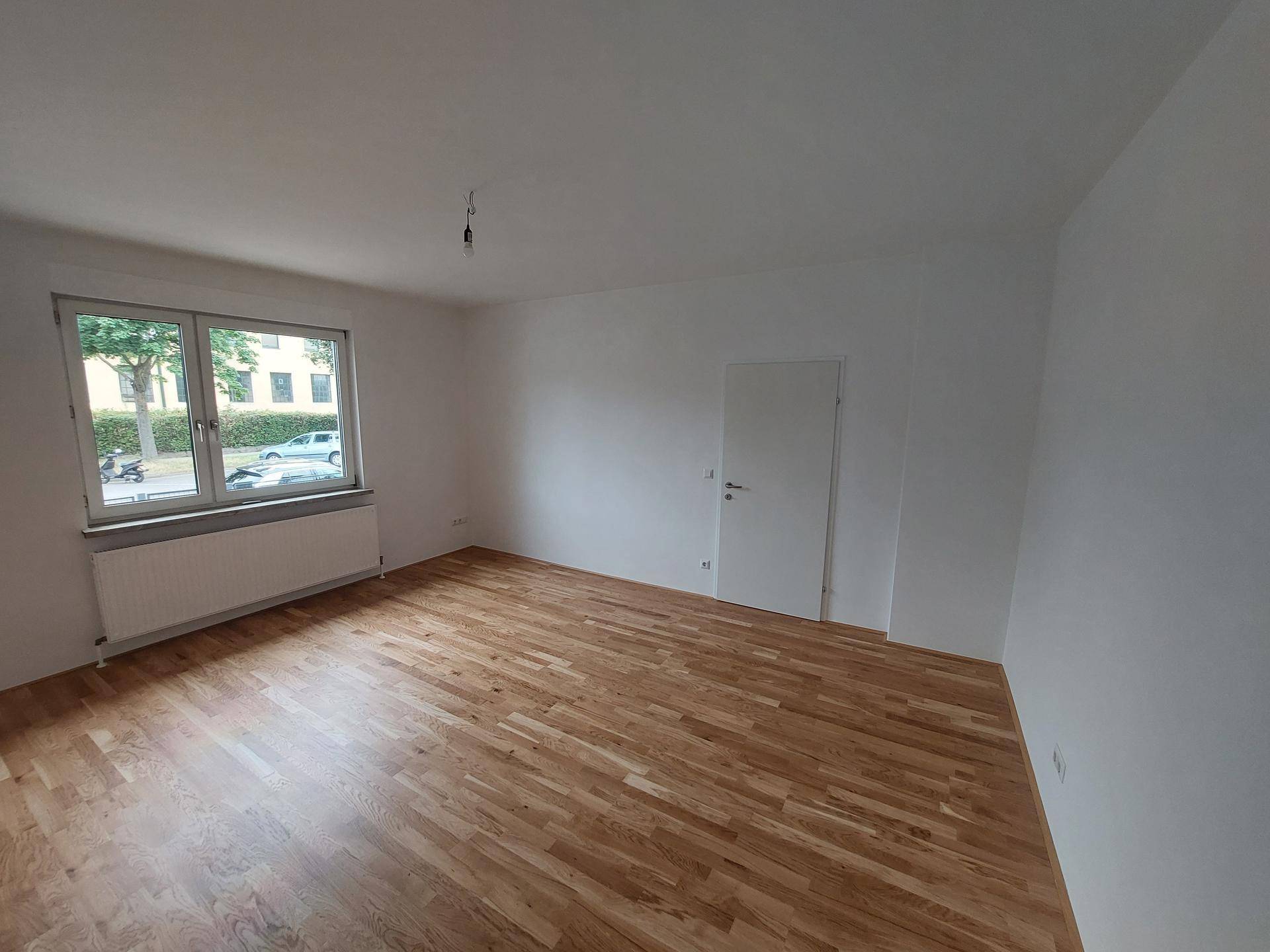 Leeres Wohnzimmer mit Holzboden, weißen Wänden und einem großen Fenster.