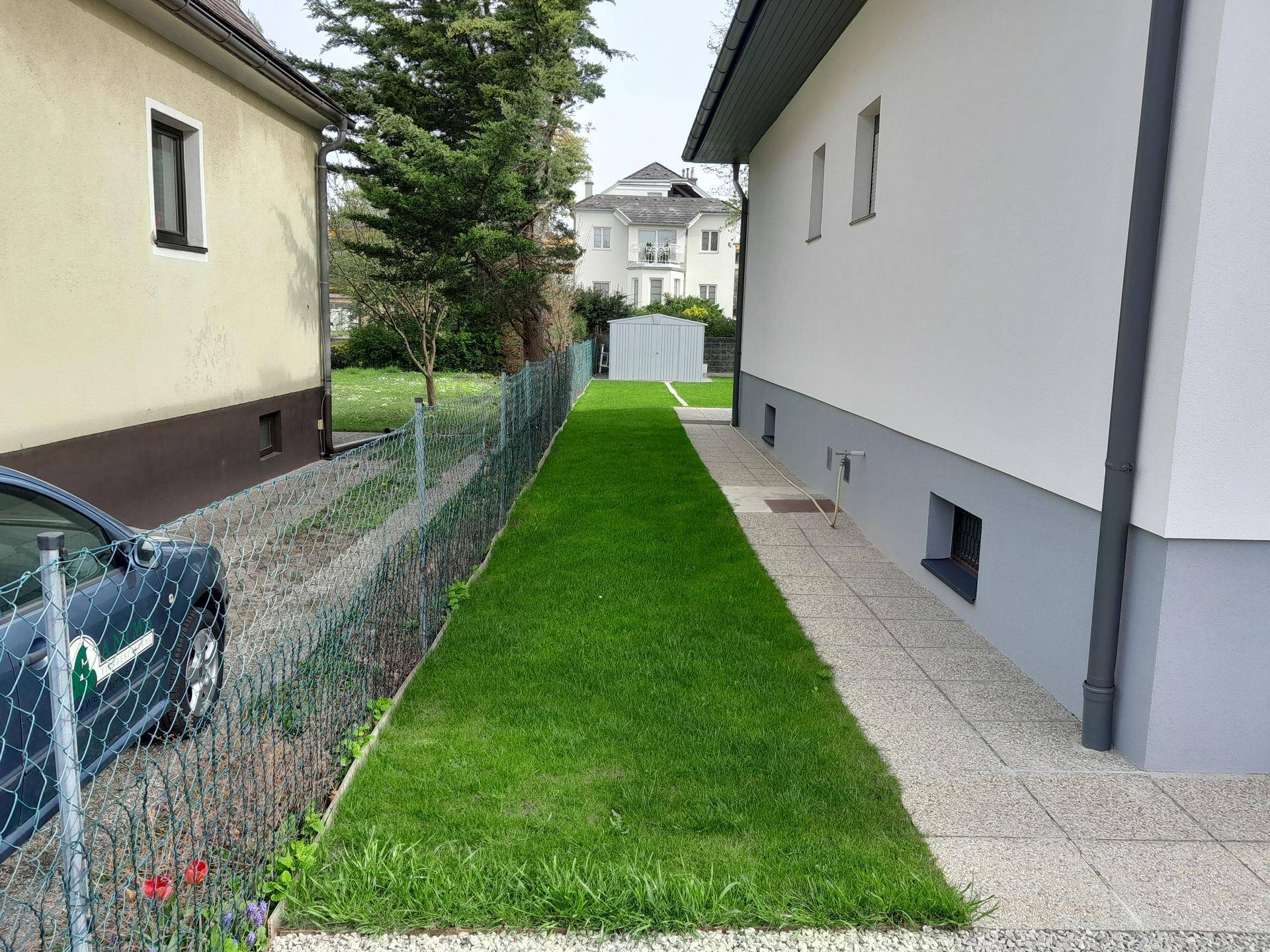 Langer, schmaler Rasenbereich neben dem Haus, umzäunt von einem Maschendrahtzaun.
