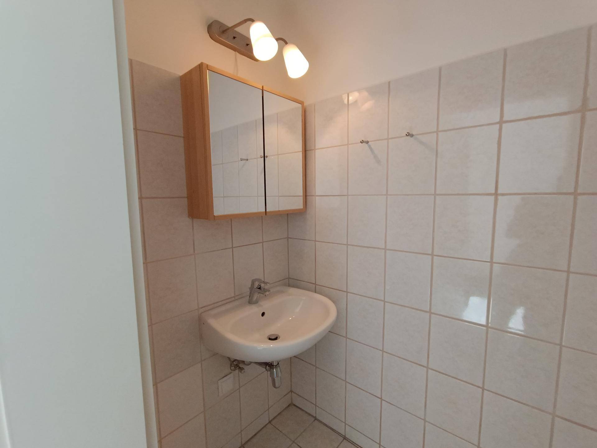 Kleines Badezimmer mit weißen Fliesen, Waschbecken und einem Spiegelschrank, funktional eingerichtet.