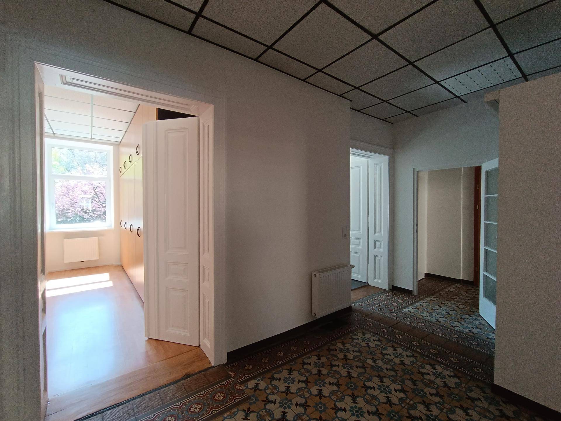 Flur mit gemusterten Fliesen, weißen Wänden und einer Decke mit Gitterstruktur, verbindet mehrere Räume.