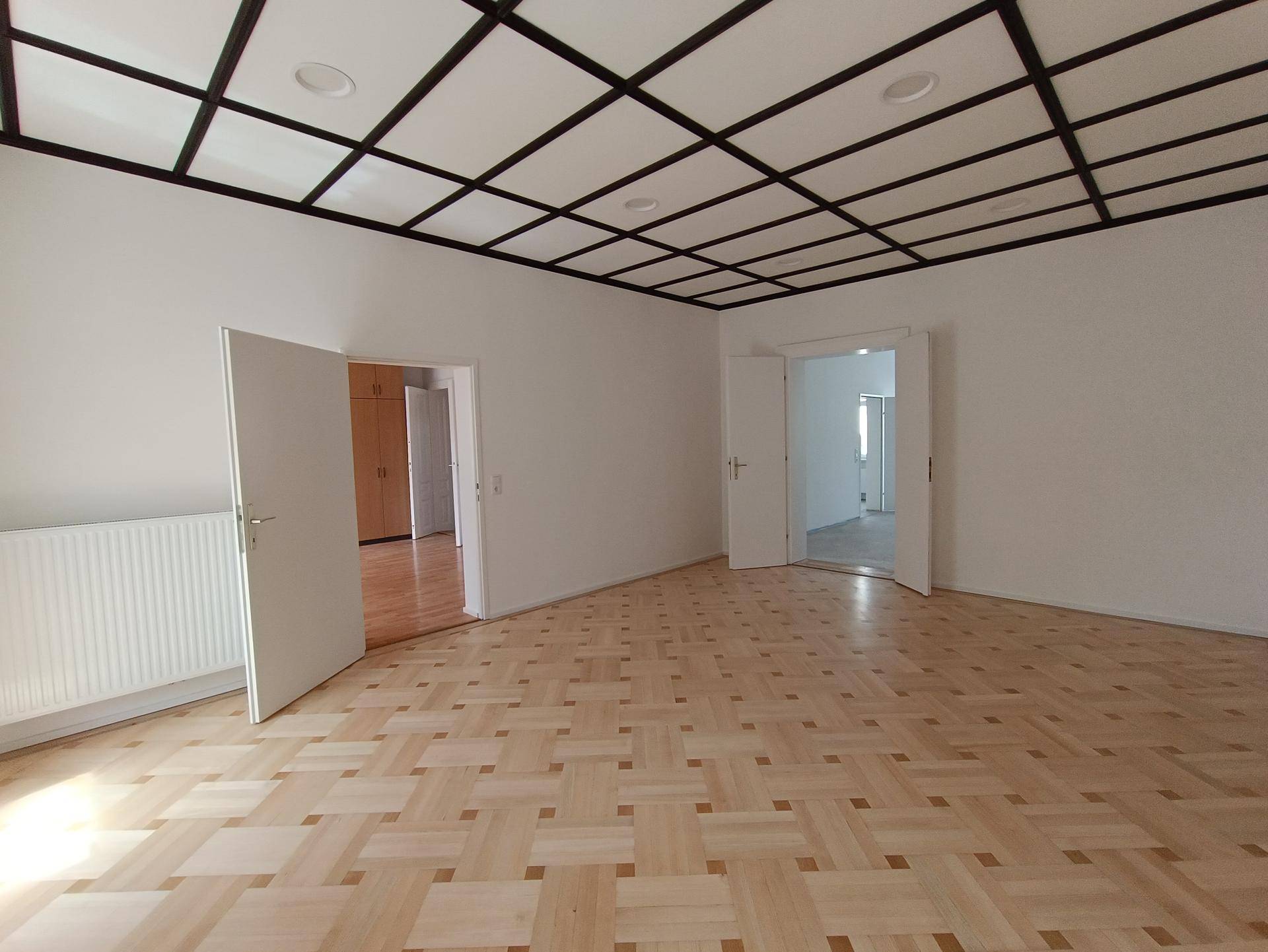 Leerer Raum mit hellem Parkettboden, weißen Wänden und einer Decke mit Gittermuster.