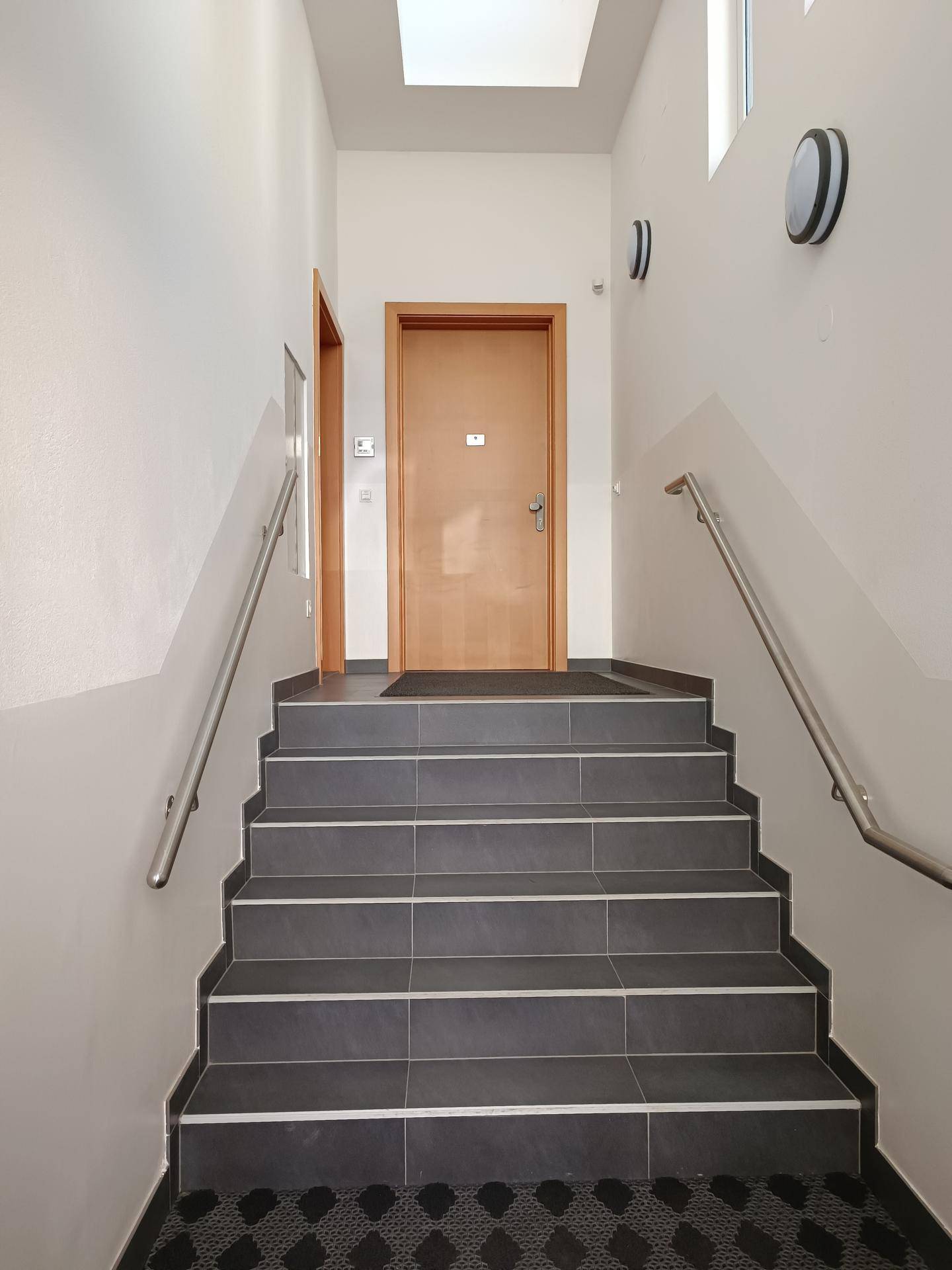 Treppenhaus mit dunklen Fliesenstufen und Metallgeländern, führt zu einer Holztür im Obergeschoss.