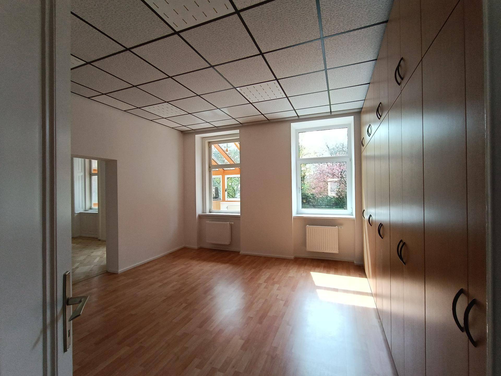 Helles Zimmer mit Laminatboden, Einbauschränken und Blick ins Grüne durch zwei Fenster.