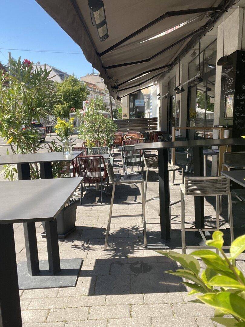 Sonnige Außenterrasse eines Cafés mit Tischen, Stühlen und Markise, umgeben von Pflanzen und Stadtblick.