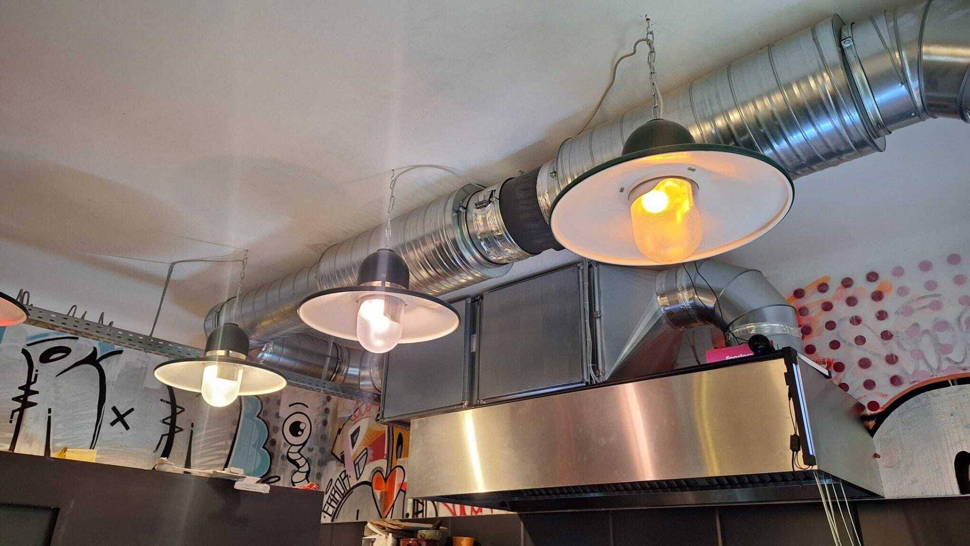 Industrielle Küche mit sichtbaren Lüftungsrohren, Pendelleuchten und einer großen Dunstabzugshaube.
