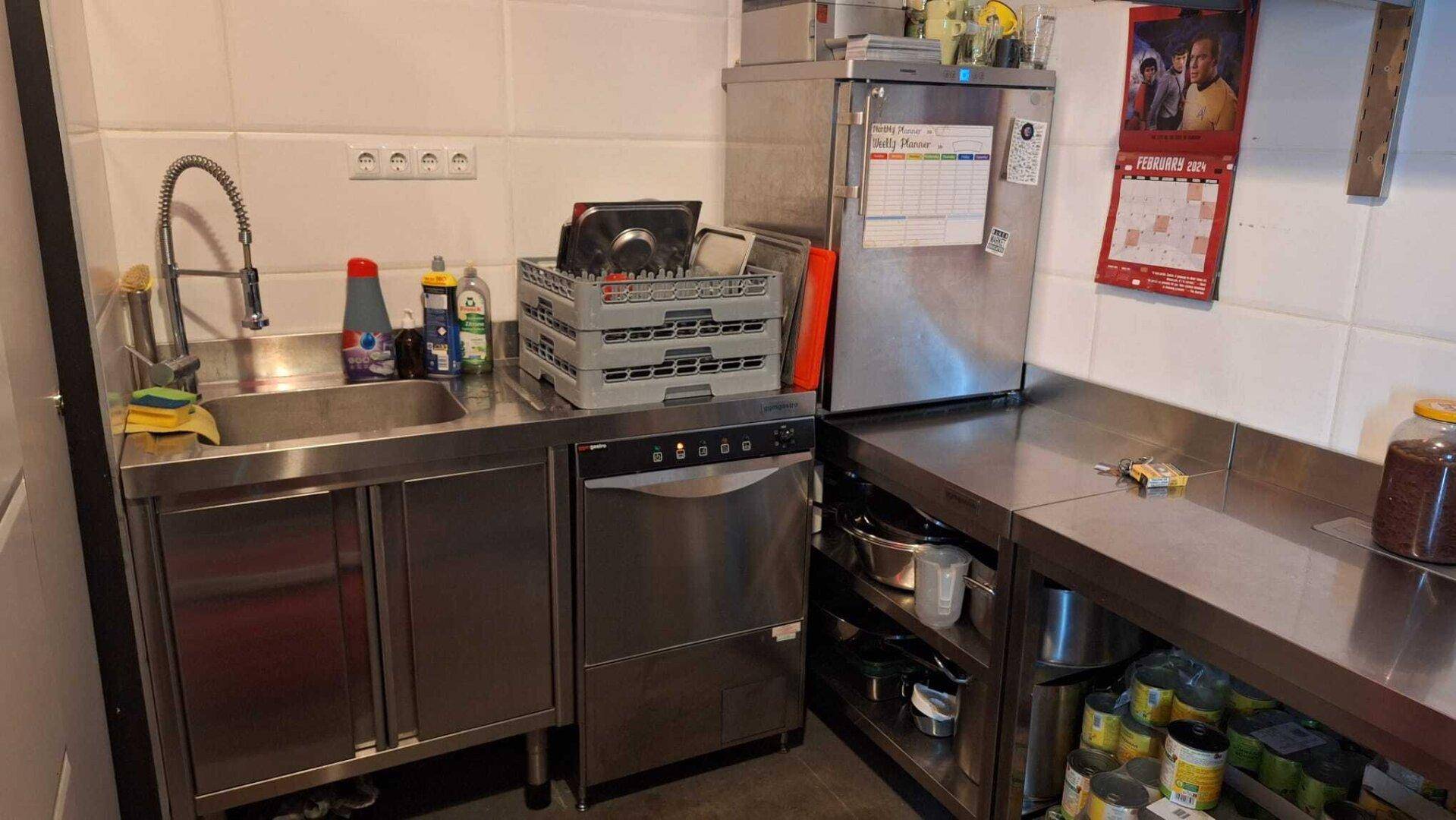 Funktionale Gewerbeküche mit Edelstahlspüle, Geschirrspüler, Kühlschrank und weißen Fliesen an den Wänden.