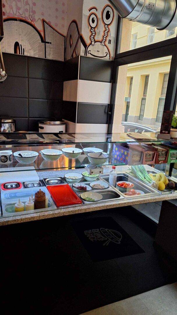 Serviertheke mit frischen Zutaten in Schalen hinter einer Glasvitrine und Wandgraffiti im Hintergrund.