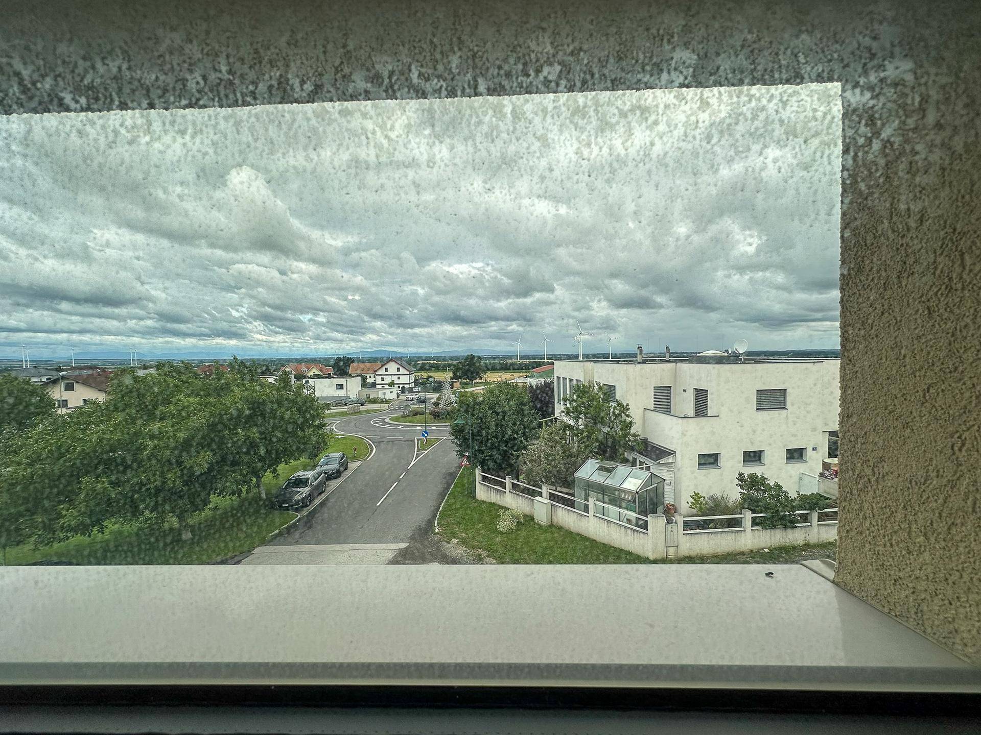 Weitblick aus dem Fenster über eine Wohnsiedlung mit Windrädern am Horizont unter bewölktem Himmel.