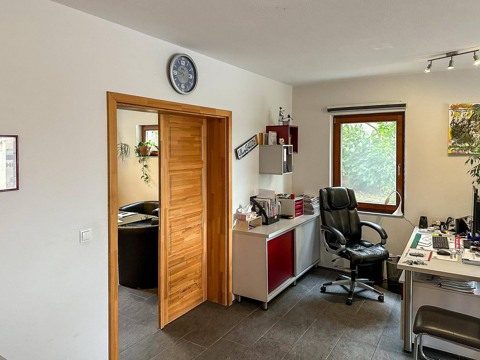 Helles Büro mit Schreibtisch, Bürostuhl und Blick ins Grüne durch große Fenster.