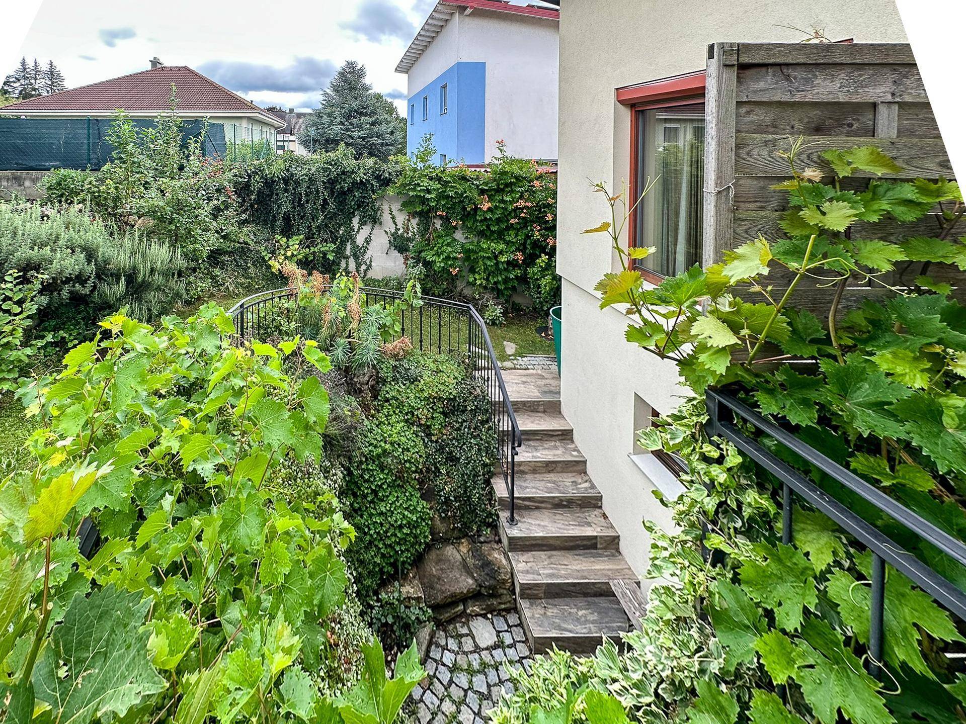 Außentreppe führt hinunter in den grünen Gartenbereich mit üppiger Vegetation und Steinpflaster.