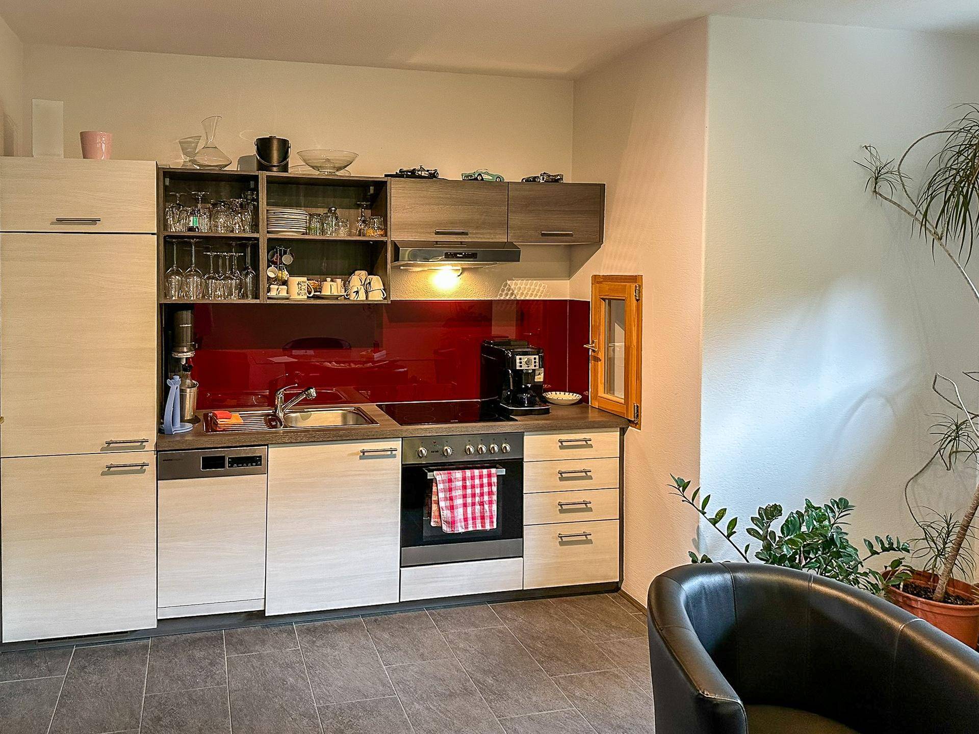 Kompakte Küchenzeile mit hellen Holzfronten, roter Rückwand und integrierten Geräten in einem Wohnraum.