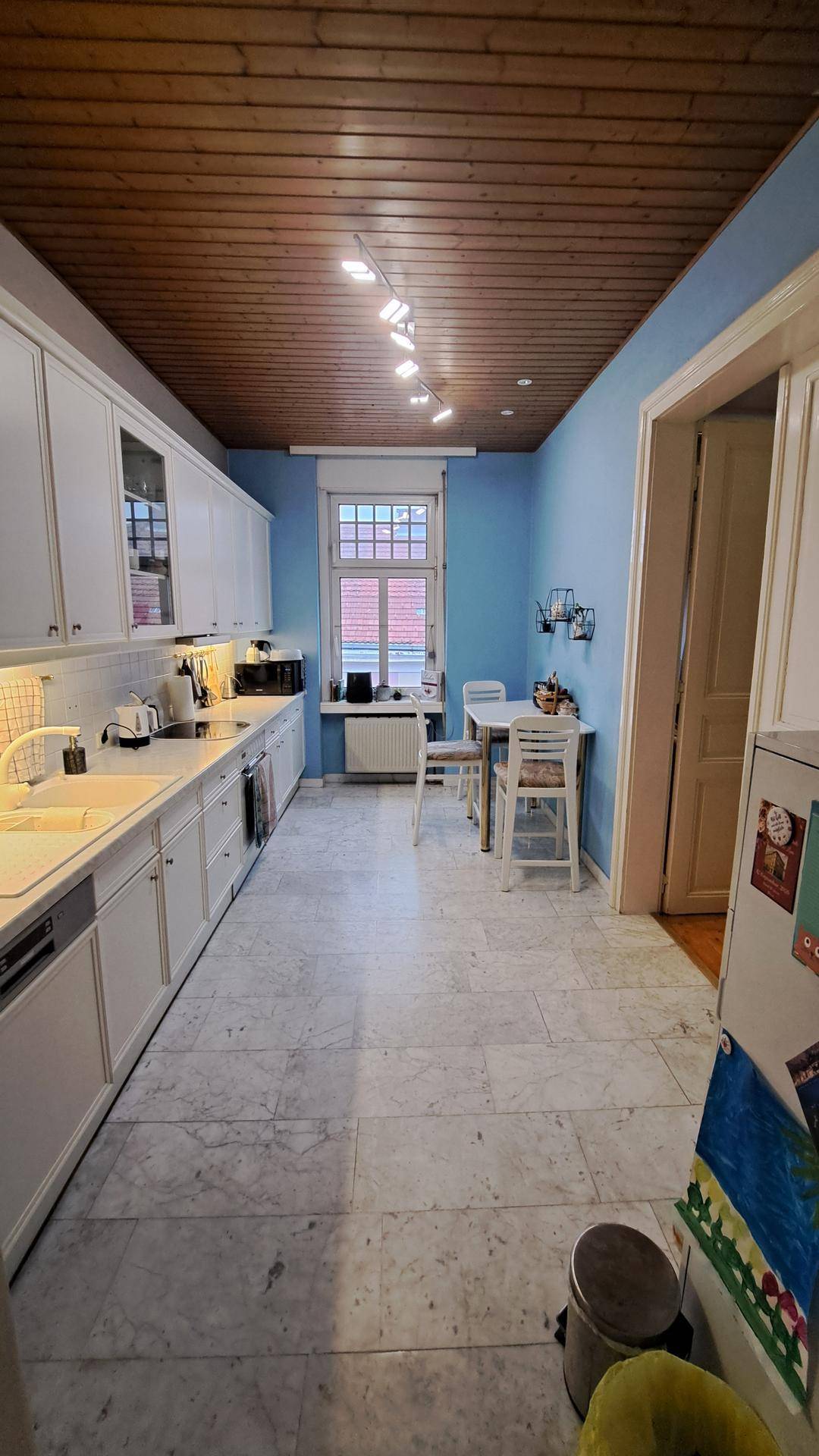Funktionale Küche mit weißen Einbauschränken, blauen Wänden und einem kleinen Essbereich am Fenster.