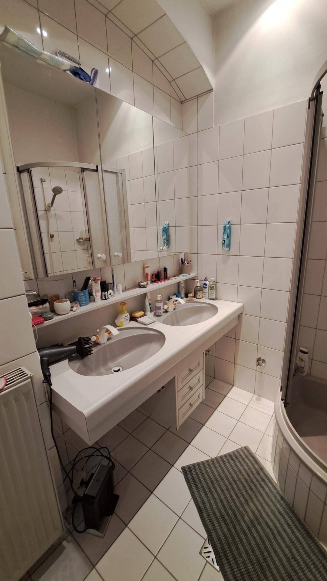 Helles Badezimmer mit weißen Fliesen, Doppelwaschbecken und einer Duschkabine, funktional eingerichtet.