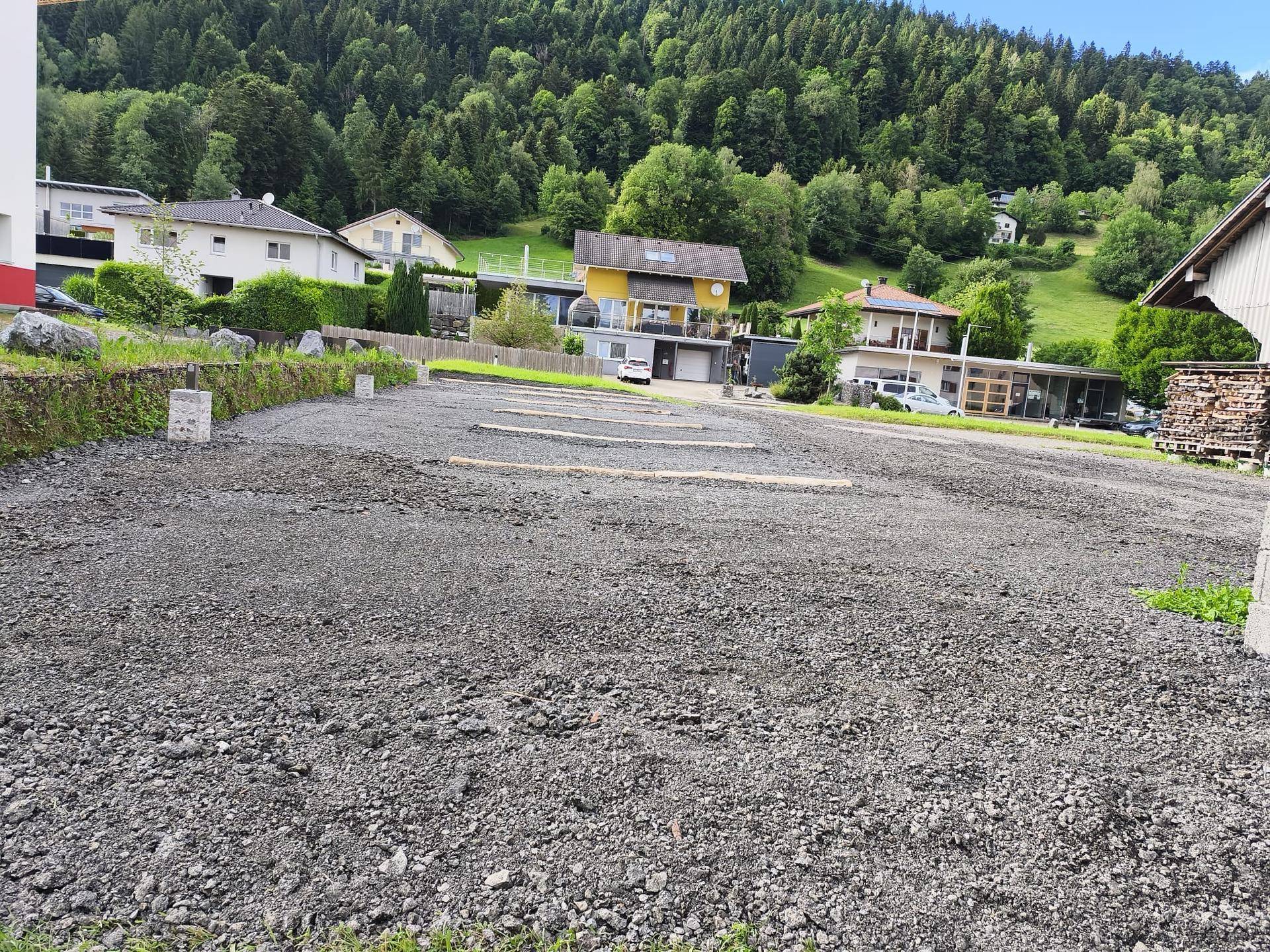 Weitläufiger Schotterparkplatz in ländlicher Umgebung mit Blick auf bewaldete Berge und weitere Gebäude.