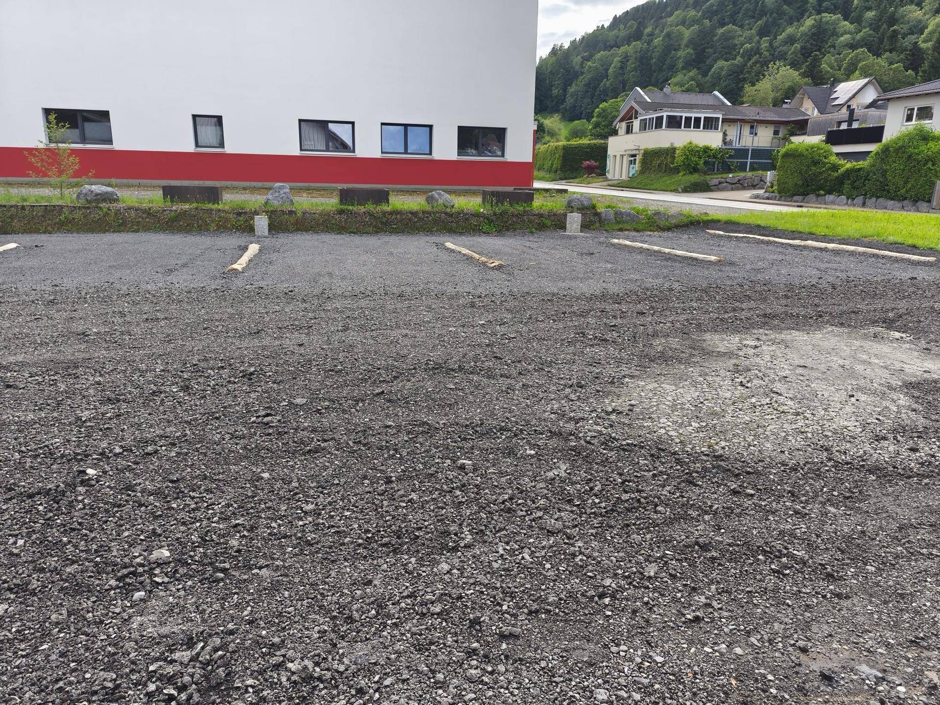 Ein im Bau befindlicher Schotterparkplatz neben einem modernen Gebäude mit roten Akzenten und Fenstern.