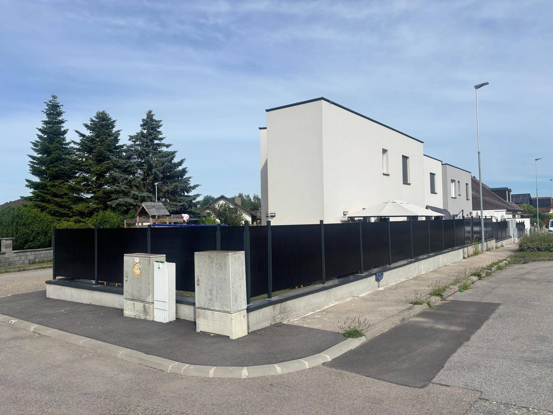 Seitenansicht des modernen Hauses mit dunklem Zaun und hohen Nadelbäumen im Hintergrund.