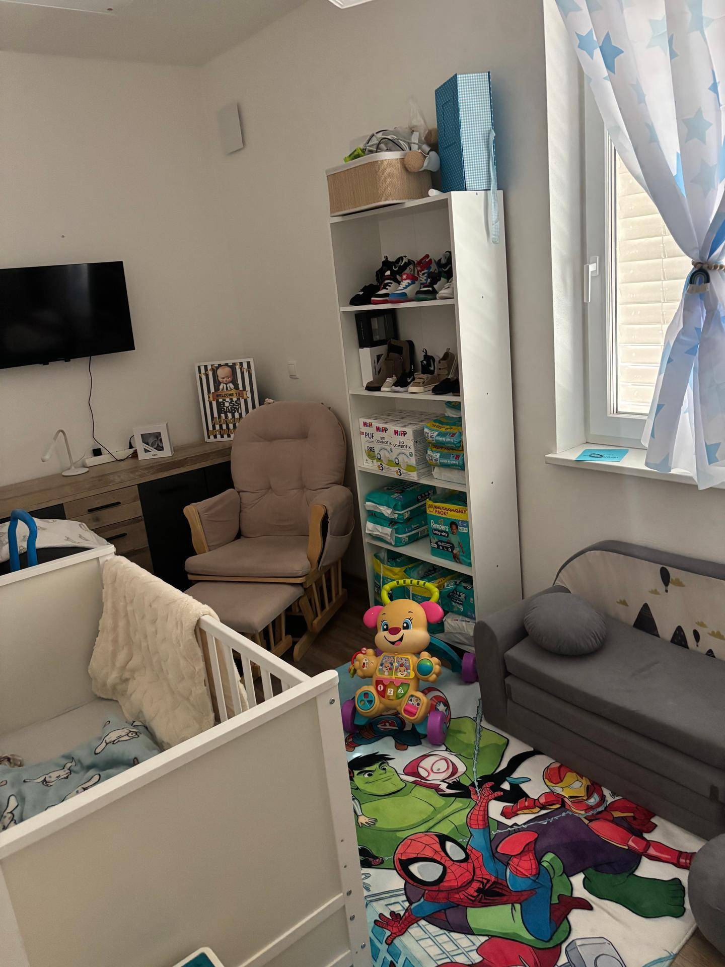 Helles Kinderzimmer mit Babybett, Schaukelstuhl, Spielteppich und Fenster.